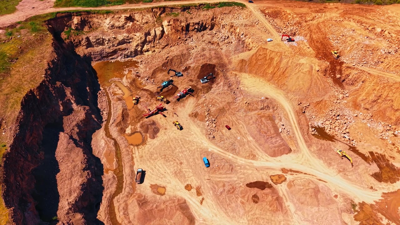High angle view of open pit mine with surrounding rock walls. Machinery and vehicles extract materials inside large gravel quarry during bright daylight.