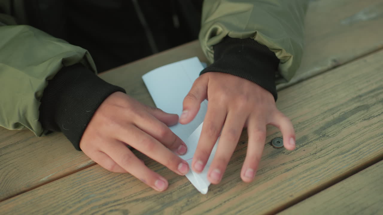 Close up of hands folding paper kite on wooden table outdoors, person wearing green jacket carefully creases edges of paper with focus and precision while sitting outside