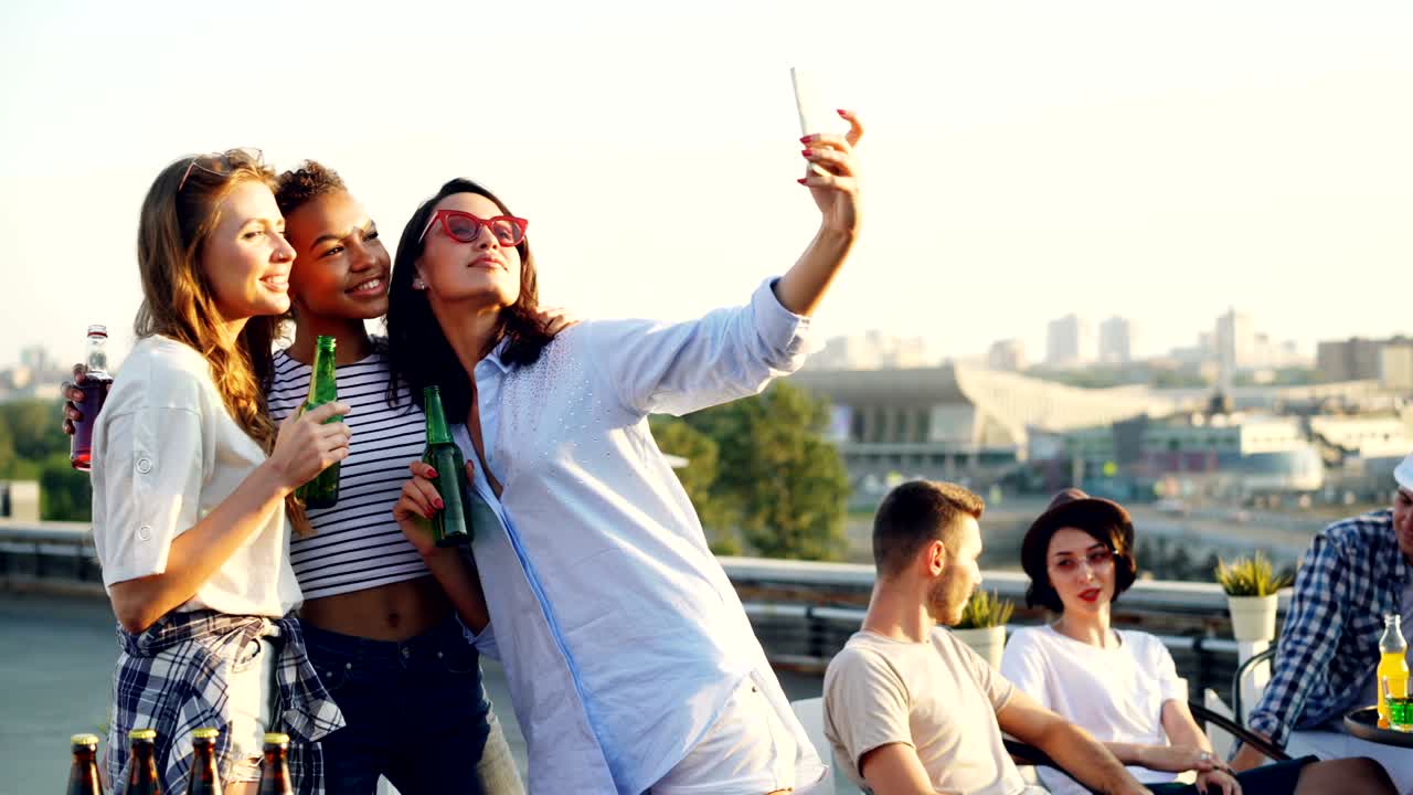 예쁜 젊은 여자들이 지붕에서 파티하는 동안 스마트폰을 사용하여 병과 함께 셀피를 찍고 친구들이 배경에서 채팅하고 있습니다.