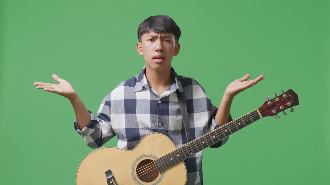 기타를 들고 있는 아시아 청소년이 당황스러운 몸짓을 하고 녹색 스크린 배경에 뭐라고 말하고 있습니다.