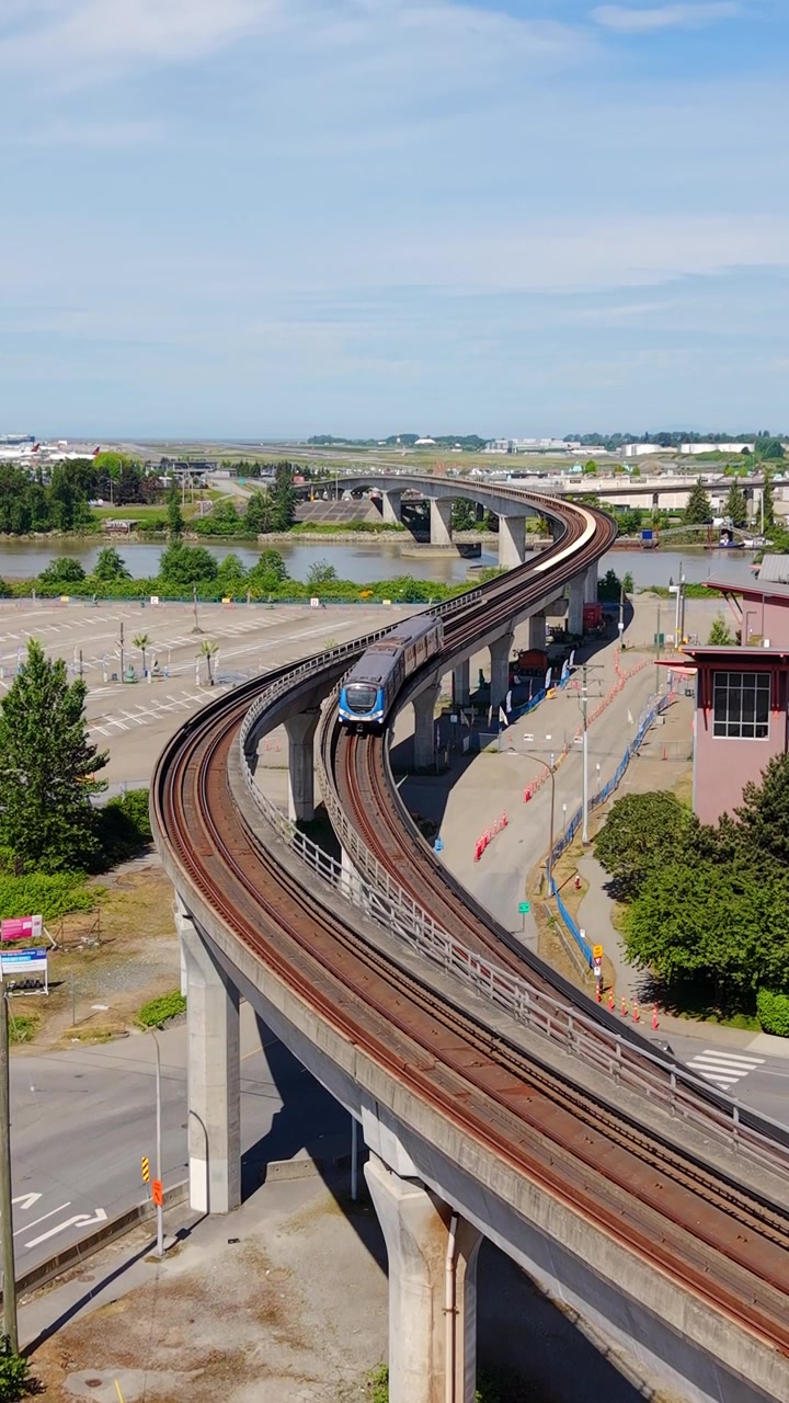 Pystykuva – SkyTrain liikkuu korotetuilla raiteilla lähellä Fraser-jokea Richmondissa, Brittiläisessä Kolumbiassa, Kanadassa. Nouseva dronekuva.