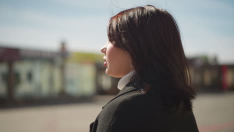 close-up of woman turning her head, brown hair swaying gently in movement, looking around with thoughtful expression in urban outdoor setting, blurred background featuring buildings