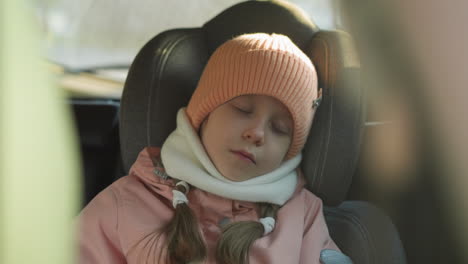 a young girl in a pink knit hat and scarf is peacefully sleeping in a car seat. the serene scene captures a moment of tranquility during a car ride on a cold day