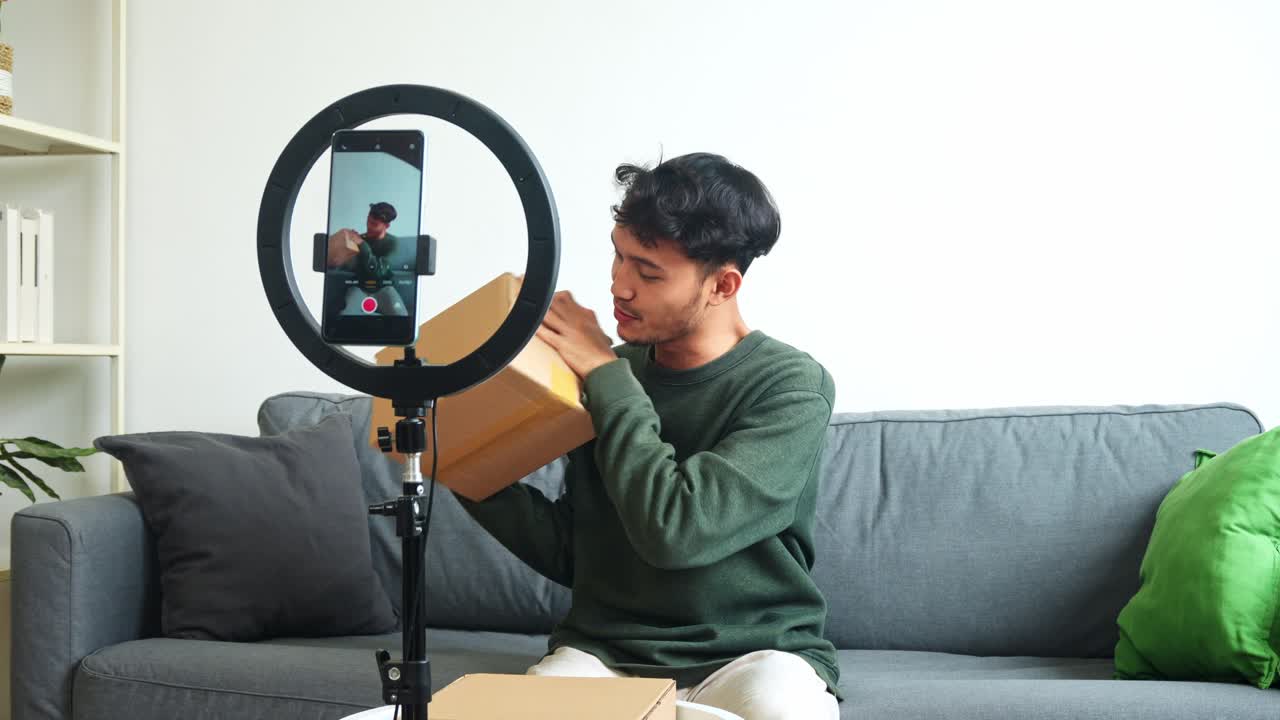 Friendly Young Asian Man Showing Cardboard To The Camera Of Smartphone While Recording Unboxing Video With Ring Light