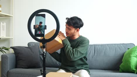 friendly young asian man showing cardboard to the camera of smartphone while recording unboxing video with ring light