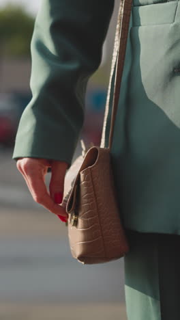 woman in green business pantsuit takes mobile phone out of leather handbag walking on street in city. successful businesswoman solves work issues at distance closeup