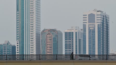 4k:-People-crossing-the-bridge,-city-traffic-and-modern-residential-towers-in-the-background,-Sharjah,-UAE