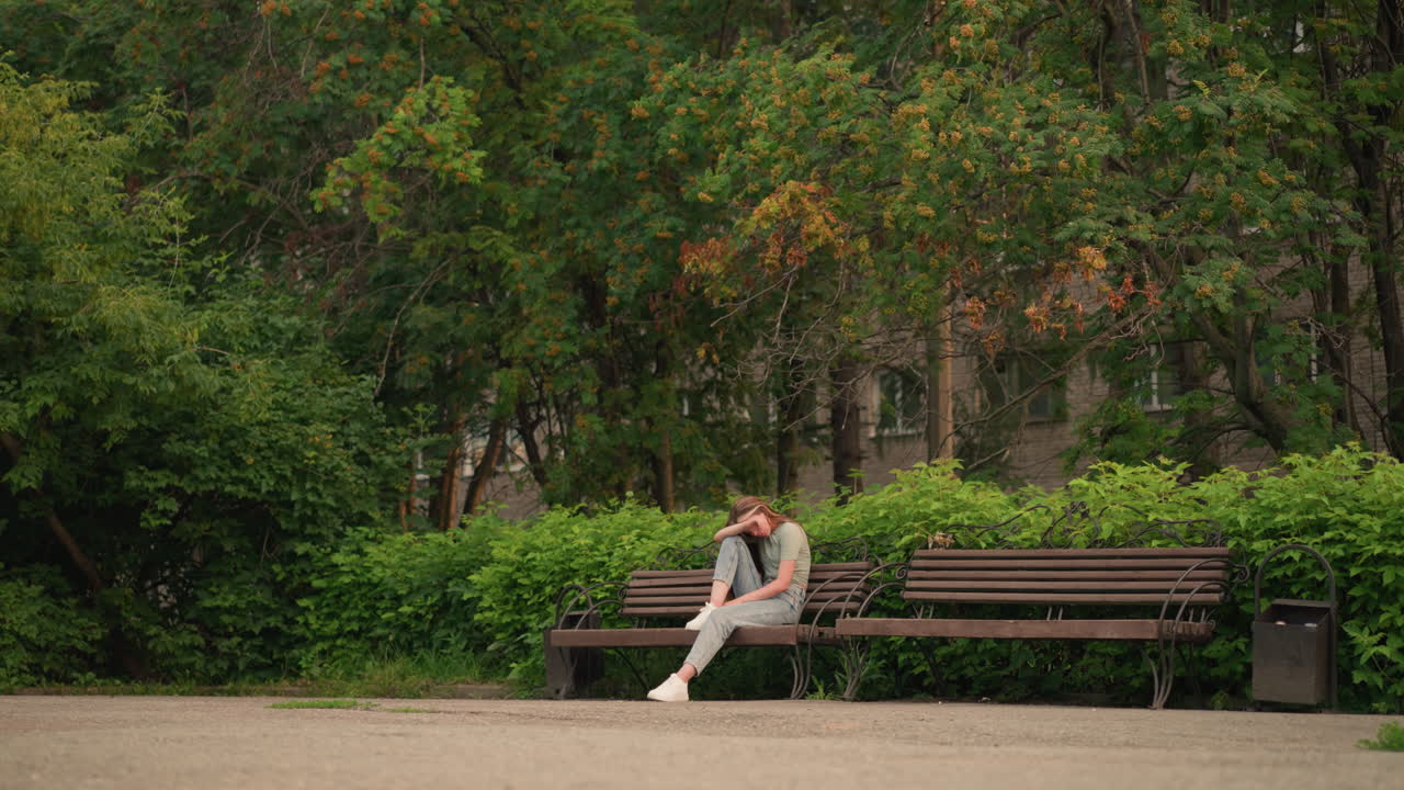 젊은 여성은 야외의 나무 벤치에 앉아, 평화로운 공원 환경에서 울창한 초록색과 나무로 둘러싸인 반사적이고 우울한 자세로 팔에 머리를 고 있습니다.