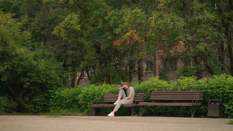 young woman sits on wooden bench outdoors, resting head on arm in reflective, somber posture, surrounded by lush greenery and trees in peaceful park setting