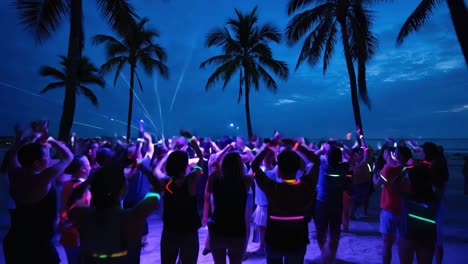 nighttime tropical beach scene featuring tourists dancing with glowing sticks, surrounded by palm trees and vibrant laser beams creating energetic party atmosphere