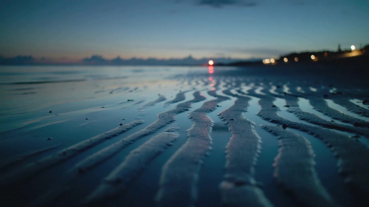 Marea crescente che spinge uno strato sottile d'acqua attraverso la sabbia increspata in una piana di marea, mostrando il riflesso di un faro rosso.