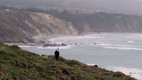 Mujer-épica-Vestida-De-Invierno-Caminando-Hacia-El-Borde-Del-Acantilado-Con-Una-Vista-Espectacular-De-La-Costa-Norte-Española