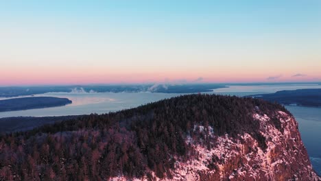 aerial view flying further away from the peak of a cliff faced mountain while seeing the misty freezing lake behind it during a winter sunrise