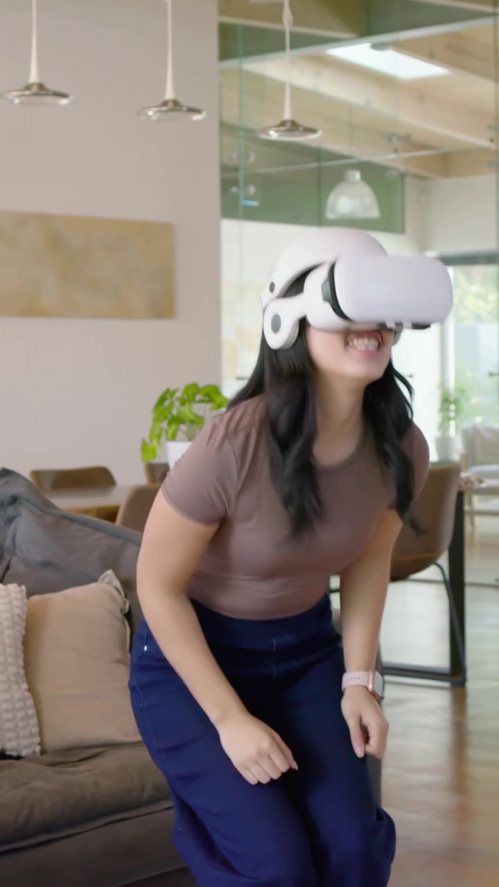 Video verticale: Donna con una maglia marrone e pantaloni blu che testa la VR a casa usando un visore bianco.