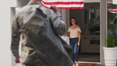 Kaukasischer-Männlicher-Soldat-Begrüßt-Sohn-Und-Frau-Im-Garten,-Während-Die-Amerikanische-Flagge-Vor-Dem-Haus-Hängt