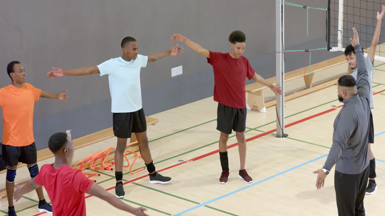 Coach signaling diverse male athletes extending arms and warming up beneath volleyball net on court