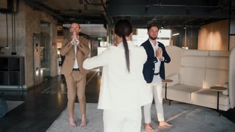 Group-yoga-class-in-a-modern-office.-A-girl-in-a-white-suit,-together-with-her-work-colleagues,-guys-in-brown-and-blue-jackets,-stand-in-a-special-pose-during-a-yoga-class-on-a-mat-in-a-modern-office