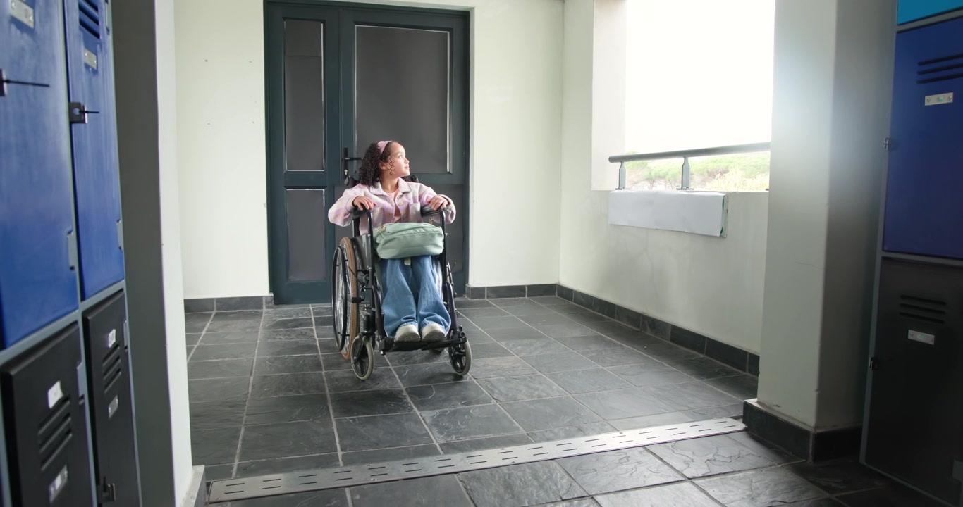Giovane donna seduta su una sedia a rotelle nella hall, con le mani sui cerchioni, che osserva il movimento fuori dalla finestra, con una borsa chiara.