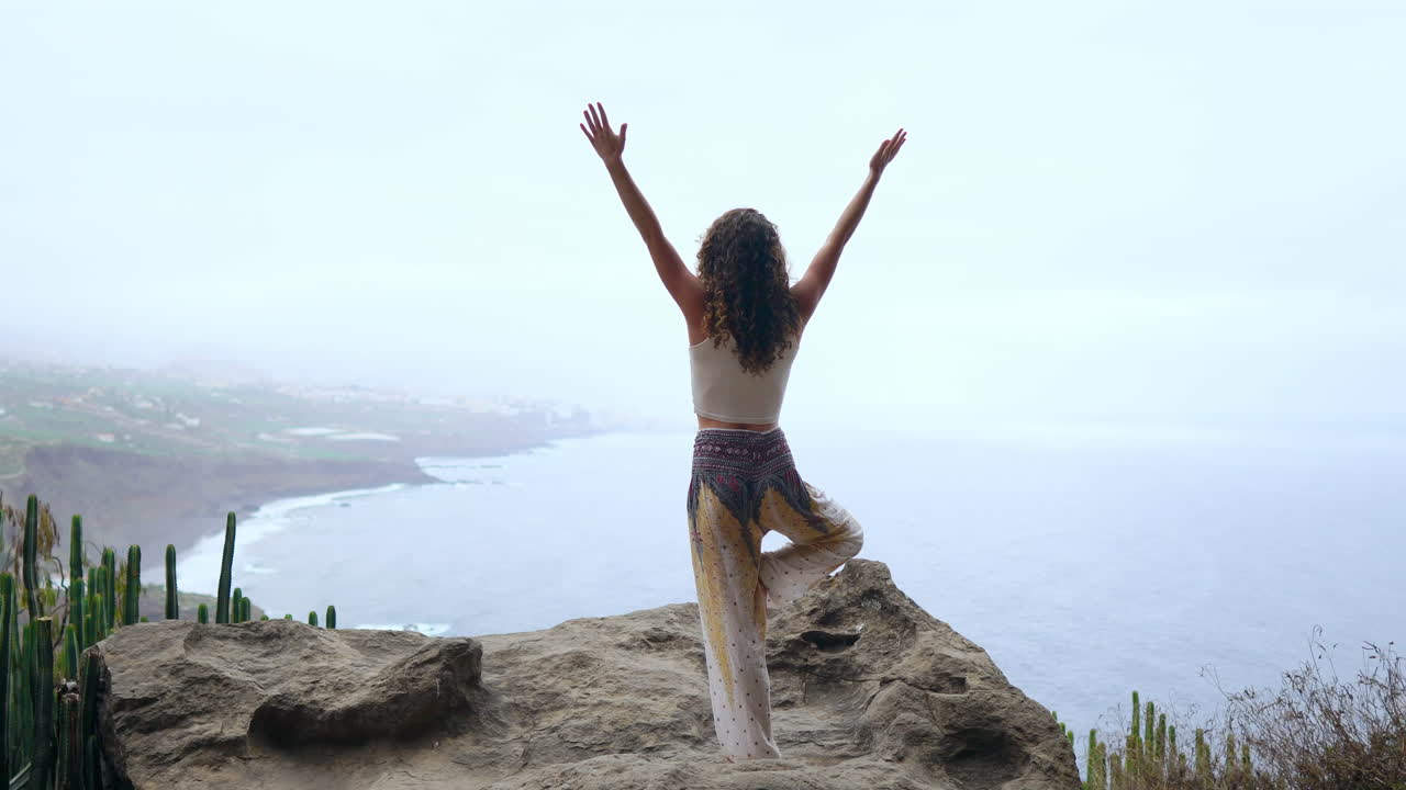 en medio de las montañas en una isla, una mujer joven hace yoga, levantando una pierna, extendiendo sus brazos, con vistas al océano