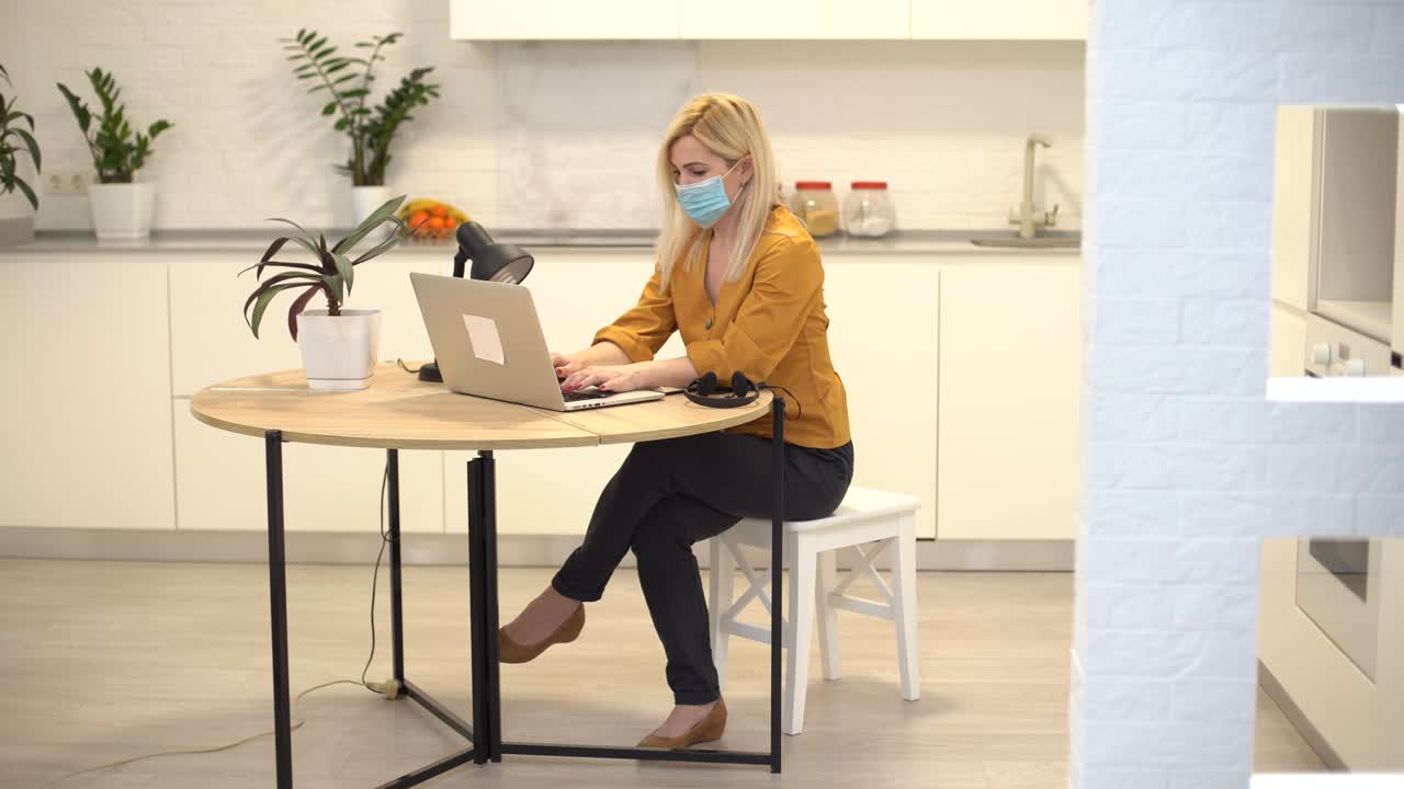 женщина в защитной маске работает удаленно на ноутбуке дома на кухне