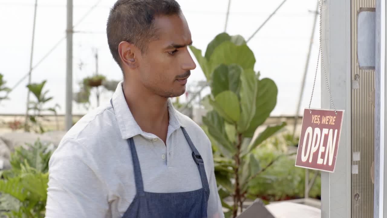 Gardener in apron standing near open sign, surrounded by lush plants, in greenhouse