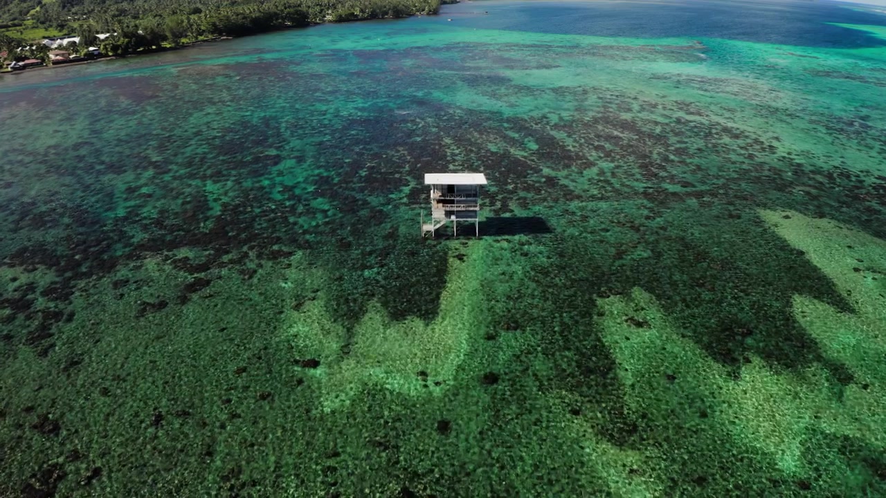 Ampia ripresa aerea con drone sopra la barriera corallina di Teahupoo, Tahiti, con la Torre dei Giudici Olimpici che si erge distante e piccola contro l'orizzonte aperto del Pacifico.