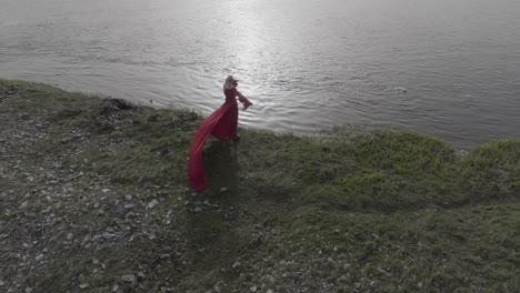 Birds-eye-drone-shot-of-female-standing-with-red-dress-at-the-shore