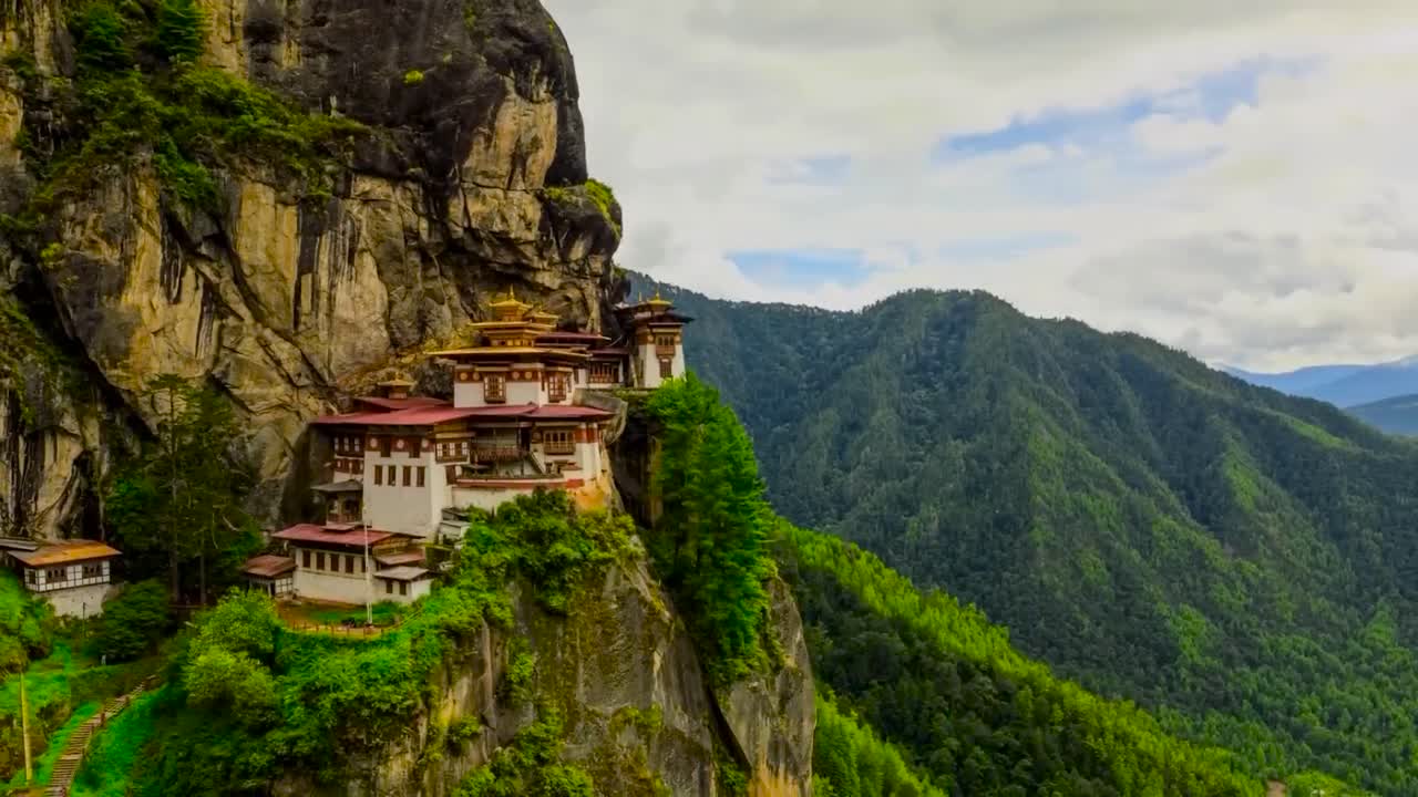 Paro Taktsang or Palphug Monastery on a steep mountain cliff during a timelapse at a sunny and cloudy day with cloud shadows running over the monastery and nature. People are seen walking in location.