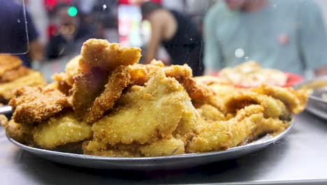 golden deep fried pork cutlets arranged on a metal tray at a bustling singapore hawker center, shot with shallow depth of field and steady camera
