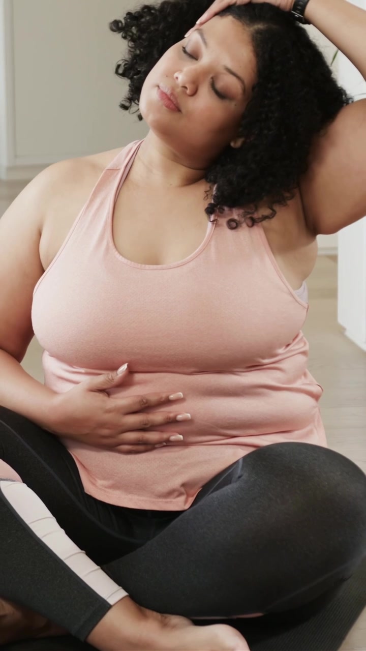 Video verticale: Una donna tocca un tablet e pratica esercizi di respirazione per la calma su un tappetino a casa, indossando una canotta rosa.
