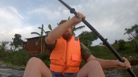 un tiro bajo de un hombre barbudo en un chaleco salvavidas remando desde las orillas del lago victoria en áfrica rural