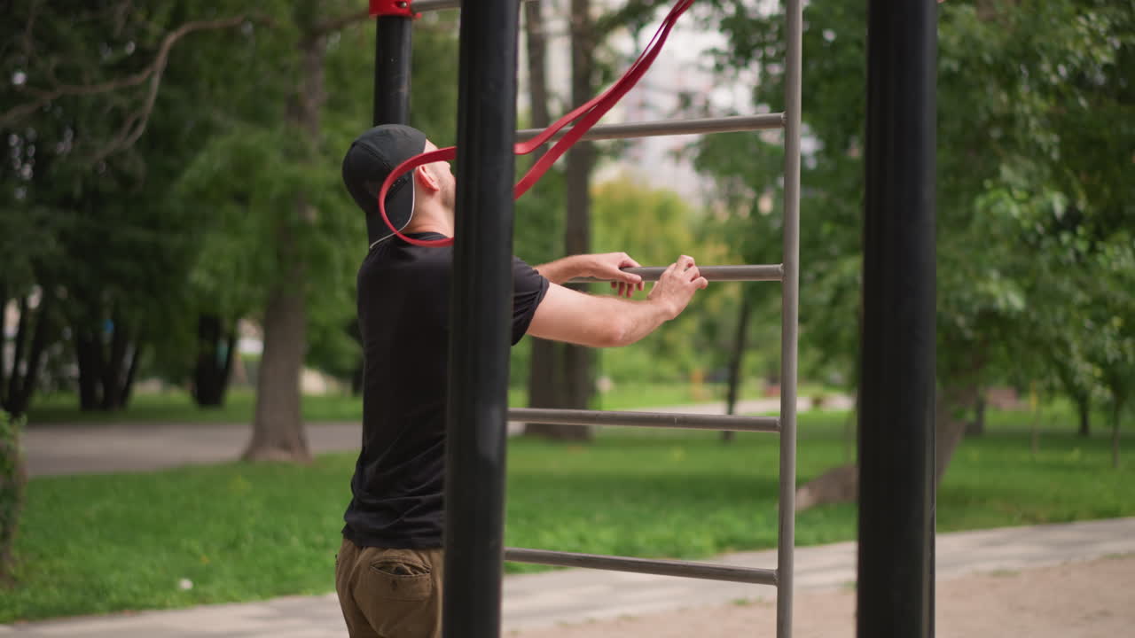 Man Gets Ready For Calisthenics Outdoors, Male Individual Secures Red Band For Outdoor Workout Session, Male Prepares His Exercise Equipment In Park Setting For Calisthenics Training