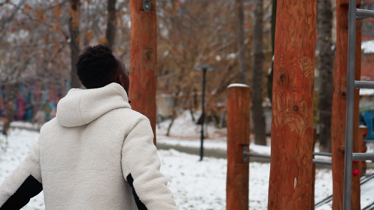 겨울 재을 입은 젊은 성인이 눈 인 공원 배경으로 야외 운동 장비의 철 바를 잡기 위해 점프하여 운동을 위해 따뜻해지는 모습