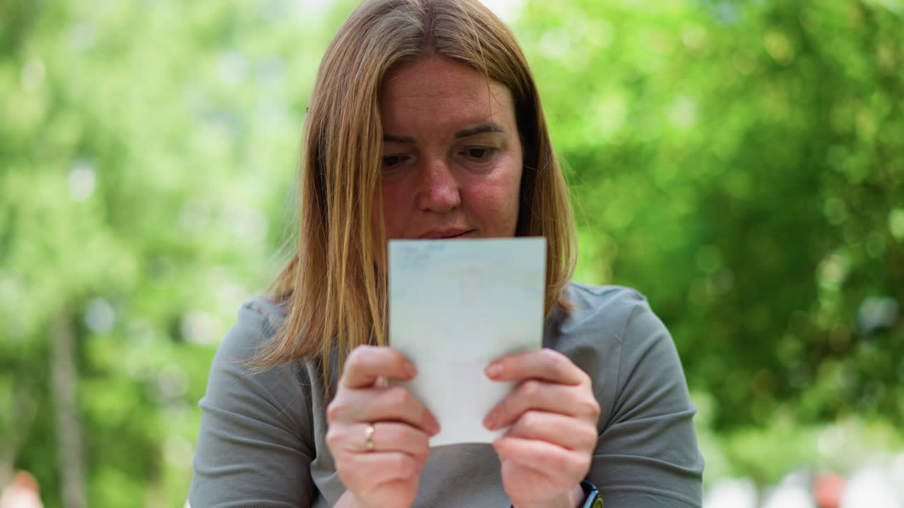 Primo piano laterale di una donna che legge un libro all'aperto e scopre un'immagine, osservando incuriosita mentre la luce del sole filtra tra le foglie verdi, in una serena atmosfera da parco immersa nella luce soffusa