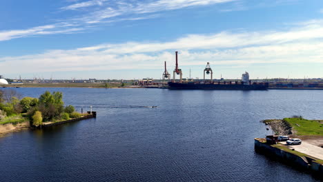 from a high overlook, a cargo ship sits at an industrial port as gantry cranes line a broad channel, calm water glittering beneath streaked clouds and quiet, efficient motion