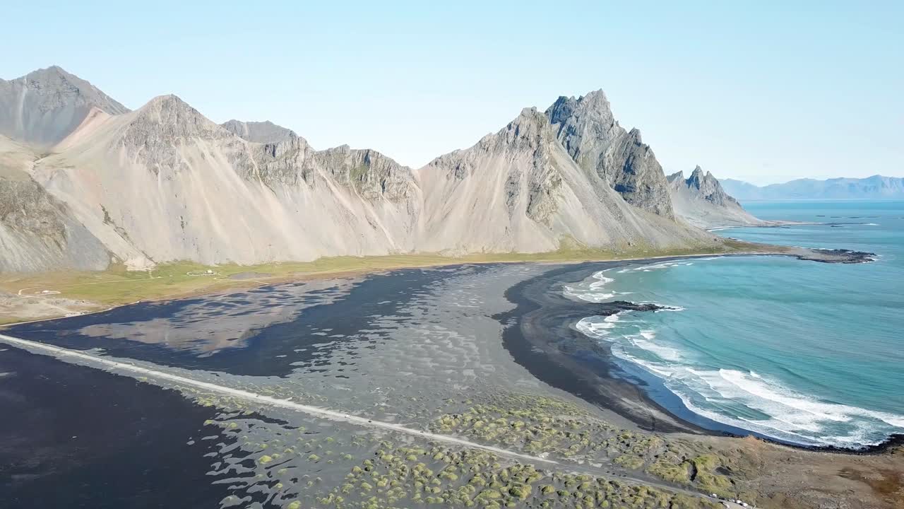 Aerial view capturing Vestrahorn mountain reflecting in the turquoise water, with waves crashing on Stokksnes black sand beach, creating a stunning scene on a sunny day in Iceland, drone panning shot