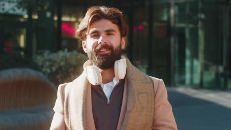 indian young businessman taking off wireless headphones looking at camera and smiling on city street