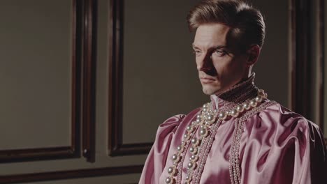 nobleman wearing pearl necklace and pink satin shirt standing confidently in vintage renaissance era interior with wooden paneled walls, embodying aristocratic elegance and historical fashion