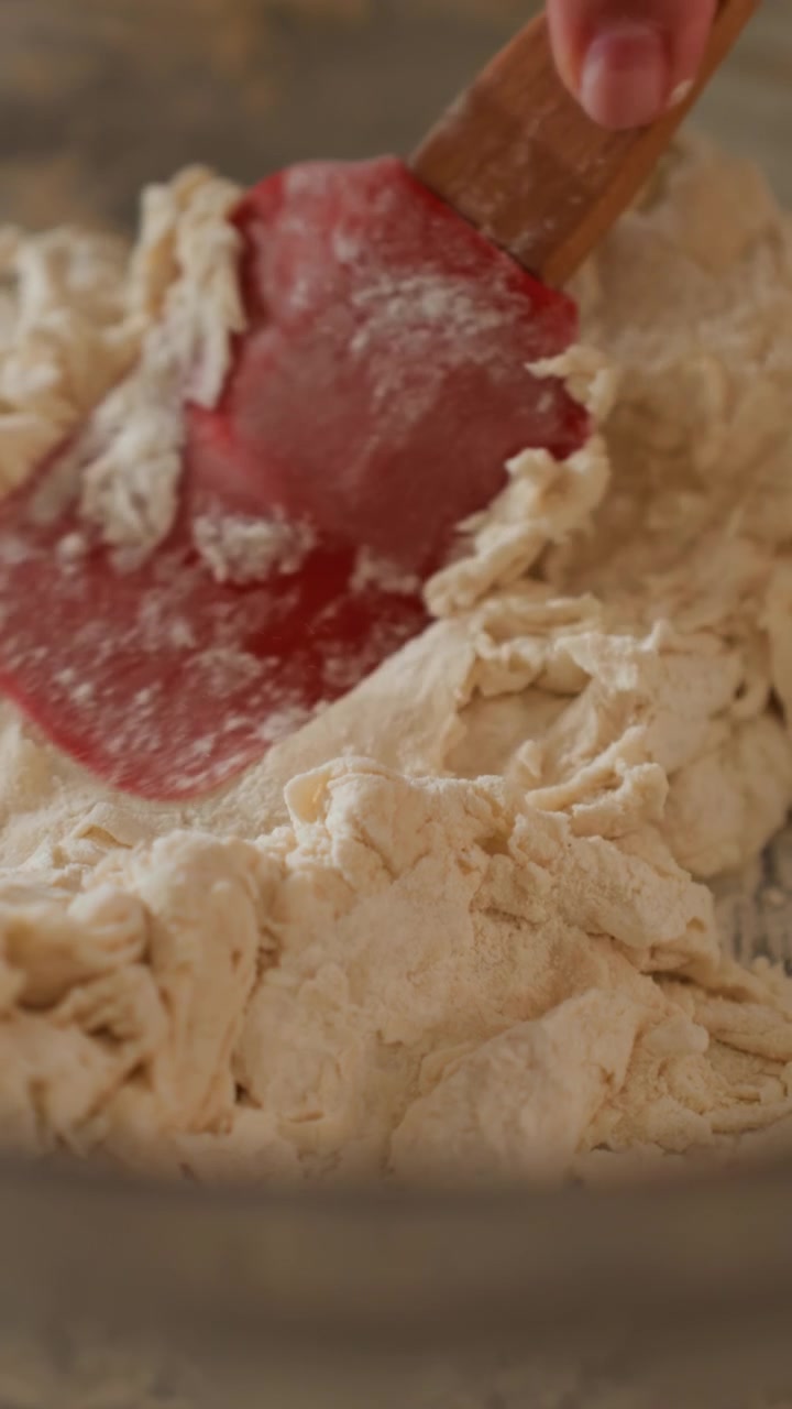 Sticky dough scraped and folded inside mixing bowl by red silicone spatula, with torn peaks, smeared ridges, flour residue, warm soft light, shallow focus, rustic kitchen prep scene