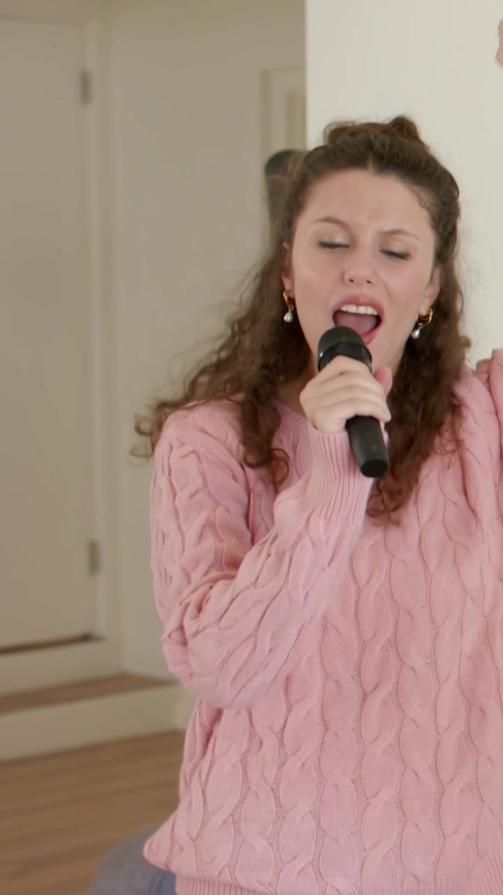 Vertical video: Hearing music, singing woman in pink sweater performing at home with black mic