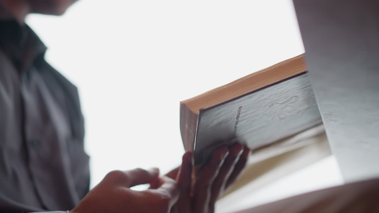 Close-up of man's hands gently flipping page of thick hardcover book near reflective table surface in bright natural light, suggesting peaceful moment of reading, focus, learning, or quiet self-reflection
