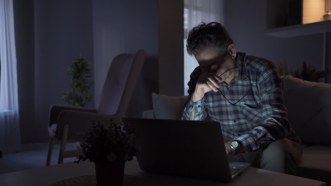 Mature man has sore eyes and sleepiness from working on laptop at night at home.