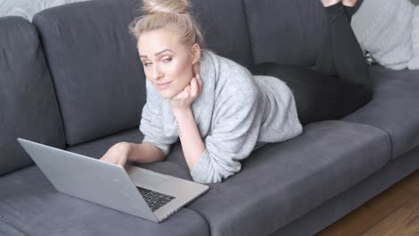 happy blond woman lying prone on sofa and working on laptop computer