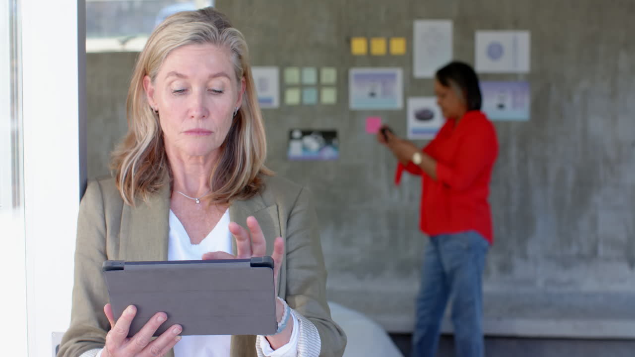 Using tablet, senior woman working while colleague in background organizing notes, in office