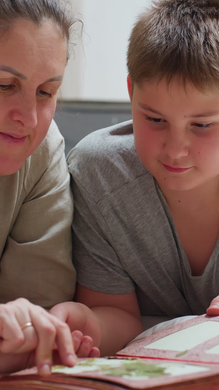 Madre e hijo blancos estudiando fotos de vacaciones en un álbum bajo luces navideñas, concentrados en las páginas, señalando con cuidado y sonriendo suavemente; la madre, como archivista familiar, preserva el legado.