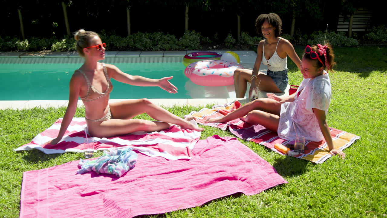 Diverse female friends relaxing by pool, enjoying sunny day with drinks and laughter