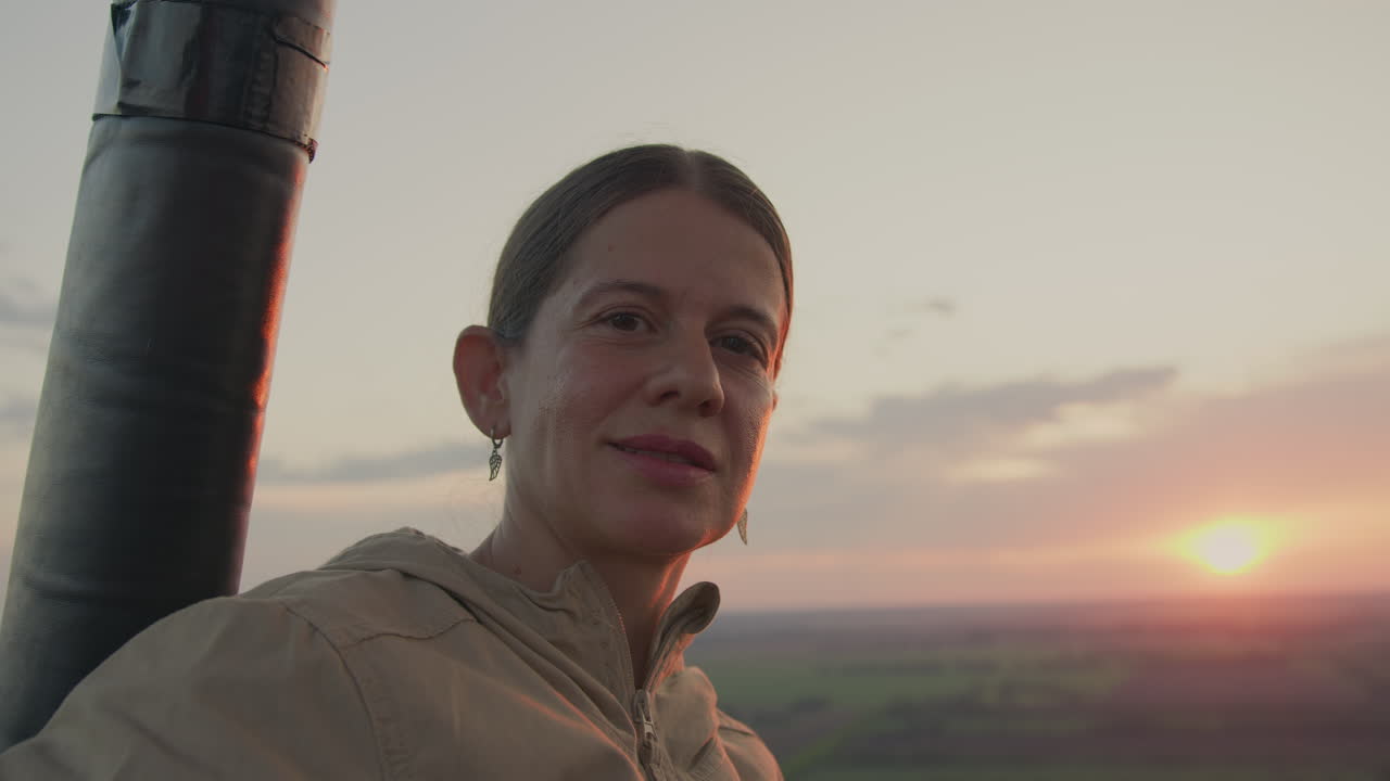 Elegant woman gazes confidently toward camera with serene smile while glowing sunset light casts soft highlights on her face, peaceful rural landscape stretching into horizon behind her