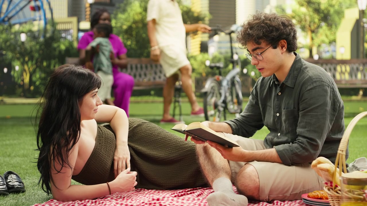 Man reading poetry book aloud to woman laying on checkered picnic blanket