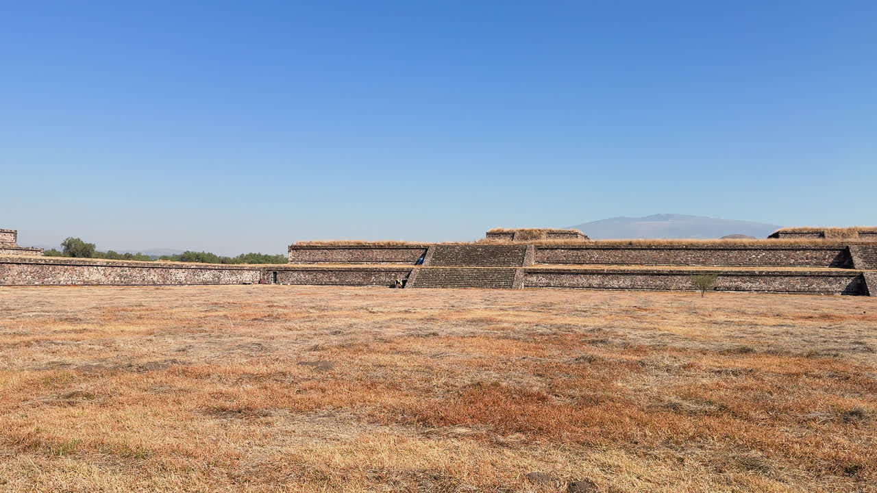 Ampia veduta delle terrazze e della piazza in pietra di Teotihuacan in Messico sotto un cielo azzurro e limpido, con erba secca in primo piano e spazio per il testo per viaggi, storia e cultura.