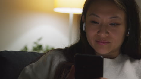 close up of woman spending evening at home on sofa wearing wireless earbuds and streaming music or podcast from mobile phone